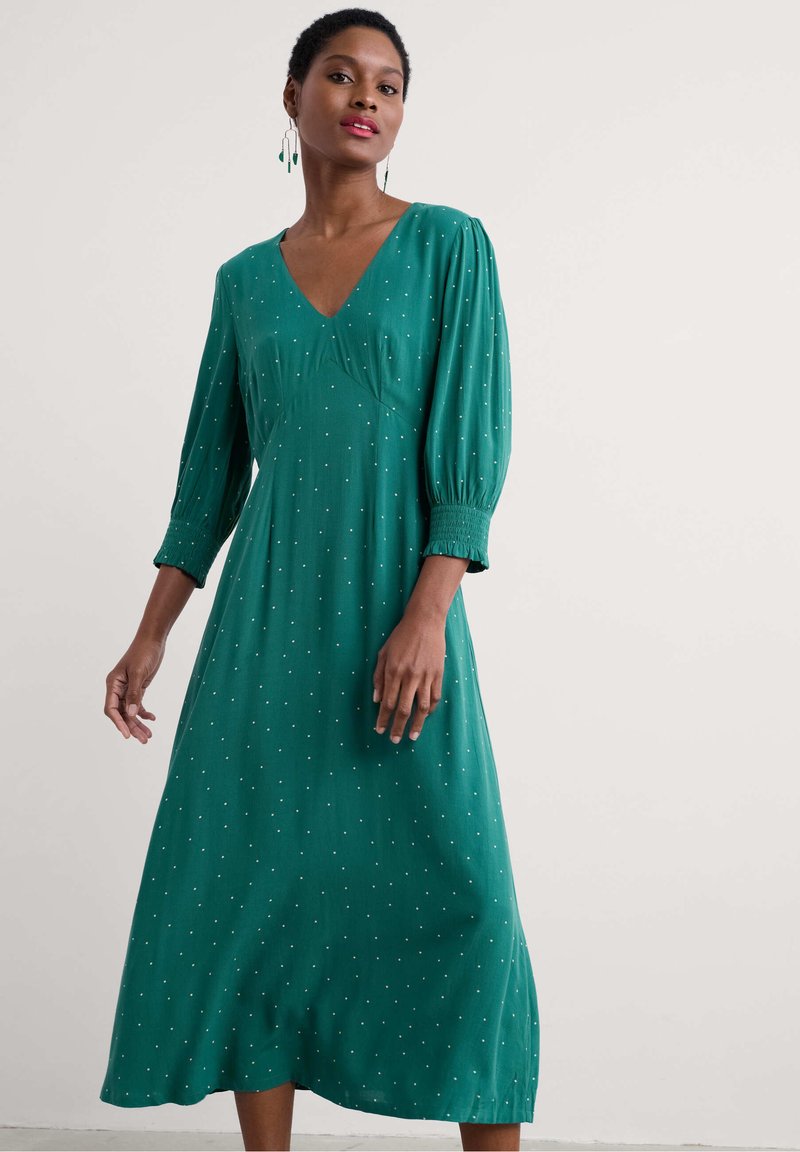 Vestido verde de manga larga con escote en V, que presenta una silueta fluida, puños fruncidos y pequeños puntos blancos dispersos por toda la prenda.