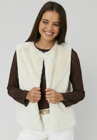 Gilet en fausse fourrure blanche avec une texture douce, design à l'avant ouvert et sans fermetures, superposé sur un top en dentelle marron à manches longues.