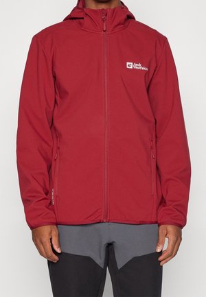 Mężczyzna w czerwonej bluzie z kapturem zapinanej na zamek, z dwoma zamykanymi na zamek bocznymi kieszeniami oraz białym logo "Jack Wolfskin" na piersi.