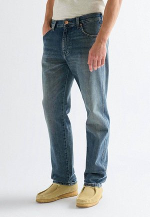 Hombre con vaqueros azules desgastados y zapatos casuales beige, de pie con una mano en el bolsillo contra un fondo liso.