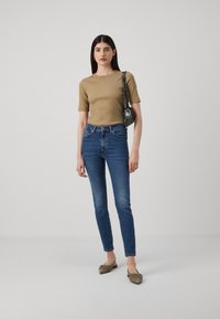 Tett khaki topp, høyt midjestrikkede blå jeans, olivengrønne flater med bue detalj, og en liten svart og grønn mønstrete håndveske. Enfarget bakgrunn.
