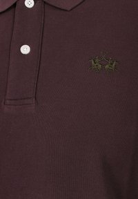 Vinröd pikétröja med texturerat tyg, tre vita knappar och en mörkgrön broderad logotyp med två ryttare.