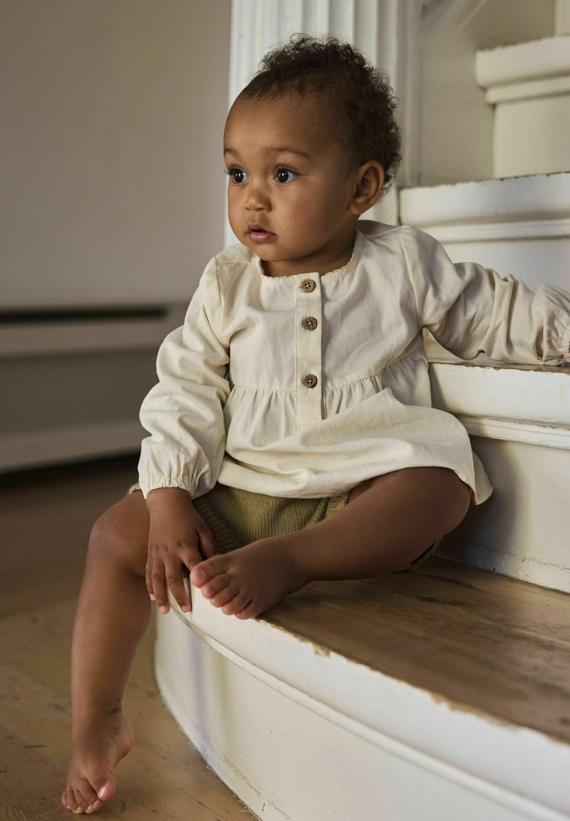 Tout-petit portant une robe crème et un short en tricot est assis sur des escaliers en bois, regardant sur le côté avec une expression calme et curieuse.