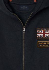 Námořnická modrá mikina na zip s označením Hackett London uvnitř límce a dvěma nášivkami na hrudi, přičemž jedna z nich zobrazuje vlajku Unie.