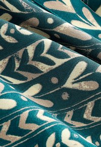 Teal tyg med ett blad- och prickmönster i kräm- och beige nyanser, med en texturerad yta och naturliga motiv som framhävs.