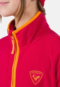 Röd fleecejacka med orange dragkedja och krage. En gyllene logotyp är synlig på vänster sida. Mjuk textur och modern design.