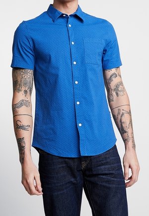 Mand iført en blå kortærmet button-up skjorte med små mønstre og mørke jeans, der viser tatoverede arme.