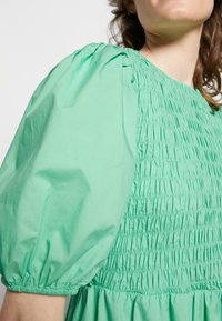 Vestido verde de algodão com mangas bufantes e corpete franzido. O tecido é suave, com uma textura franzida ao redor do decote e da cintura.