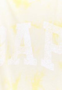 Gelbes Batik-T-Shirt mit auffälligem weißen "GAP"-Schriftzug in abgenutztem Look. Weicher Baumwollstoff, lässiger Stil, lockere Passform.