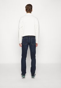 Sweatshirt branco, jeans azul-escuro e sapatilhas com detalhes verdes. Os jeans têm um corte clássico com bolsos traseiros e detalhes de costura visíveis.