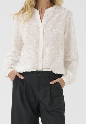 Kobieta w prześwitującej białej bluzce z kwiatową haftem i czarnych spodniach z wysokim stanem, ręce w kieszeniach.