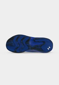 Semelle de chaussure de sport bleue et noire vue de dessous, montrant des motifs ondulés et le logo blanc Under Armour près du talon.