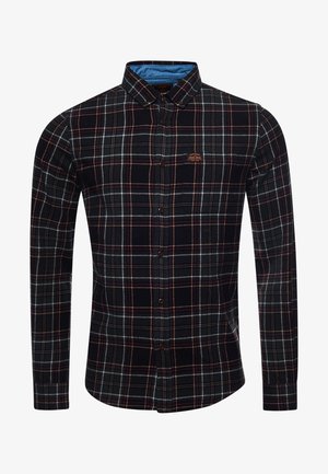 Homme portant une veste en cuir marron par-dessus une chemise à carreaux noirs et verts, jean bleu et baskets blanches, debout contre un mur en panneaux de bois.