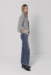 Szara kurtka bomberka na czarnym topie, w połączeniu z niebieskimi dżinsami o prostych nogawkach i czarnymi botkami. Modelka stoi na tle białego tła.