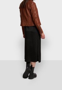 Braune Lederjacke mit tailliertem Schnitt und Reißverschlussakzenten, getragen über einem schwarzen Midikleid. Sichtbare schwarze klobige Stiefel mit strukturiertem Sohlen.