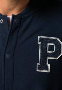Gros plan sur une personne portant une veste bleu marine avec une lettre "P" en relief blanc sur la poitrine et de sombres boutons-pression à l'avant.