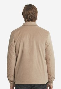 Beige cordflodjacka med långa ärmar, krage och rak passform. Har ett texturerat tyg med subtila vertikala ränder och knappade ärmslut.