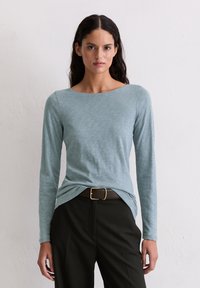 Gai zila, garām piedurknēm, apģērbs ar apaļu kakla izgriezumu, kas izceļas ar maigu tekstūru un brīvu piegriezumu, kombinēts ar melnām biksēm un brūnu jostu.