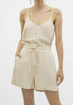 Vrouw draagt een beige mouwloze top met knopen en dunne bandjes en bijpassende hooggetailleerde short met trekkoord, staand met één hand in de zak.