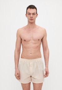Boxers en coton beige avec une taille élastique et une fermeture à bouton à l'avant, présentés sur un modèle masculin contre un fond blanc uni.