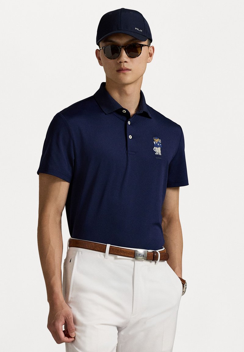 Polo shirt de culoare albastru marin, cu mâneci scurte, guler cu două nasturi și detalii brodate. Asortat cu pantaloni albi și o curea maro.