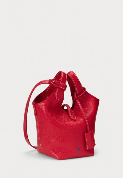 Sac seau en cuir rouge avec surface texturée, forme arrondie, double poignées et une petite étiquette. Présente des surpiqûres contrastées et un accent de logo.