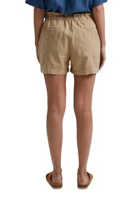 Personne portant un short beige taille haute avec des poches arrière et des sandales marron à enfiler, debout, les pieds légèrement écartés, sur fond blanc.