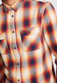 Camisa de franela a cuadros en rojo, amarillo y marino con cuello abotonado, dos bolsillos en el pecho y botones marrones. La tela parece tener textura.
