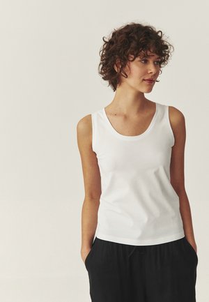 Ung person med krøllet hår iført en hvid ærmeløs top og sorte bukser, stående med hænderne i lommerne og kiggende mod højre.