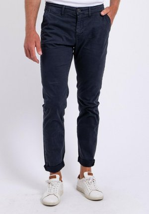 Homme portant un pantalon bleu marine foncé retroussé avec des baskets blanches, debout, une main dans la poche sur fond blanc.