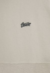 Sweat-shirt beige en tissu texturé, avec un logo foncé imprimé au centre. L'ourlet présente un détail tissé subtil.