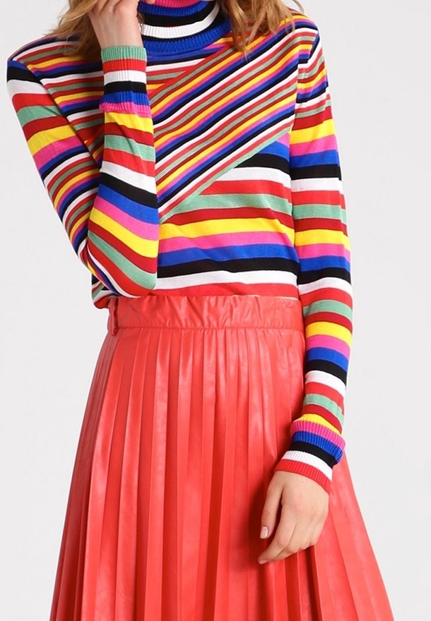 Femme portant un pull à col roulé rayé coloré associé à une jupe plissée rouge taille haute, la main reposant près de la poitrine et de la hanche.
