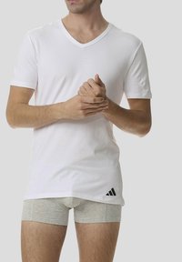 Vit bomull v-ringad t-shirt med korta ärmar, med en svart Adidas-logga i nederkant till vänster. Grå boxerkalsonger är delvis synliga.