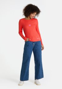 Frau trägt ein rotes langärmliges Shirt mit einem rosa Herz und blauen weiten Jeans, steht mit einer Hand in der Tasche und blickt nach unten.