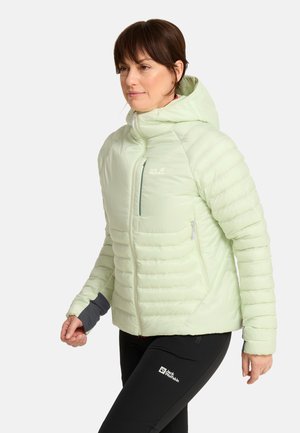 Vrouw draagt een lichtgroene gewatteerde jas met capuchon en een zwarte broek, staand en iets naar links kijkend tegen een effen achtergrond.