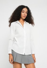 Donna con capelli ricci che indossa una camicia bianca a bottoni con colletto di gioielli e una mini gonna grigia plissettata, in piedi di fronte a uno sfondo bianco uniforme.