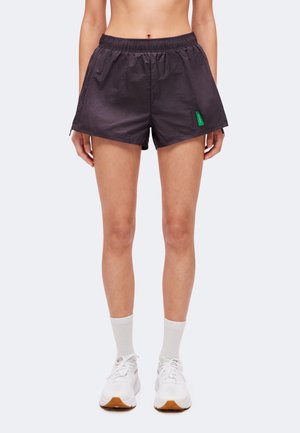 Vrouw met zwarte sportshorts met een elastische tailleband, witte sokken en witte hardloopschoenen, staand op een effen lichte achtergrond.