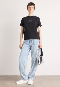 Zwarte katoen t-shirt met korte mouwen en Calvin Klein-logo. Gecombineerd met lichtblauwe wijde jeans en witte sneakers, terwijl een metalen tas wordt vastgehouden.