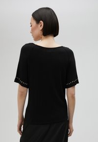 Černý top s krátkým rukávem a širokým výstřihem, který má ozdobné accenty podél rukávů, měkkou tkaninu a uvolněný střih.