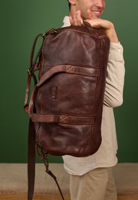 Borsa da viaggio in pelle marrone con diverse tasche, zip robuste e una tracolla staccabile. Finitura texturizzata con accenti a contrasto.