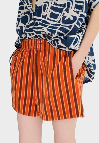 Personne portant un short rayé orange et marron avec une taille élastique et une chemise à motifs bleus et blancs, les mains dans les poches.