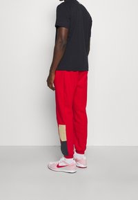Rote Sweatpants mit einem beigen Patch und marineblauem Akzent, kombiniert mit weißen und pinken Sportschuhen, vor einem schlichten Hintergrund.