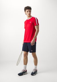 Camiseta deportiva roja con acentos blancos y logo de Fila, combinada con shorts azul marino. El modelo sostiene una raqueta de tenis, vistiendo zapatillas oscuras y calcetines blancos.