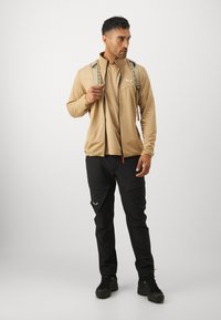 Beige zippered jakke over en tan skjorte, kombinert med svarte cargo-bukser. Antrekket har et to-tonet design og en ryggsekk med mønstrede stropper.