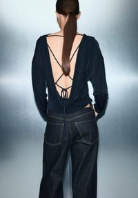 Czarna bluzka z długim rękawem i otwartym tyłem, posiadająca krzyżujące się ramiączka, w połączeniu z ciemnoniebieskimi spodniami typu wide-leg z żółtymi szwami.