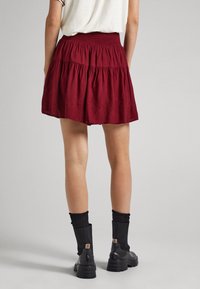 Person som bär en röd volangkjol, vit topp, svarta strumpor och svarta ankelboots som står mot en enkel grå bakgrund.