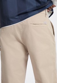 Beige cotton pants with a smooth texture, featuring a back pocket and a small branded tag near the waistline.