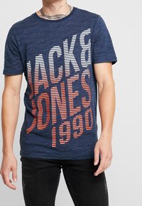 Muž v námořnicky modrém tričku s diagonálními bílými a oranžovými pruhy s textem "Jack & Jones 1990" a černých poškodených džínách.