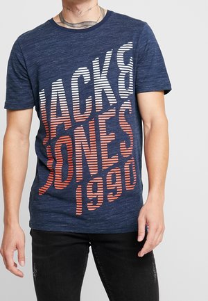 Hombre vistiendo una camiseta azul marino con texto en diagonal de rayas blancas y naranjas "Jack & Jones 1990" y jeans negros desgastados.