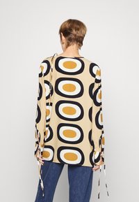 Långärmad topp i beige tyg med ett stort cirkulärt mönster i svart, vitt och senapsgul. Justerbara band vid handleden.